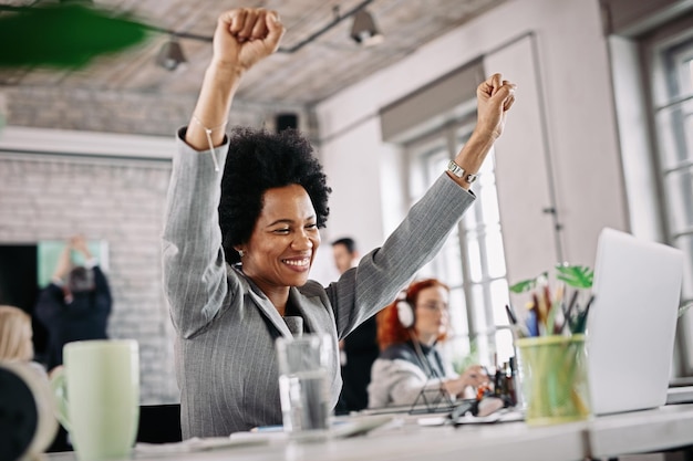 Vista de ángulo bajo de la feliz mujer de negocios negra que trabaja en la oficina y celebra su éxito con los brazos levantados