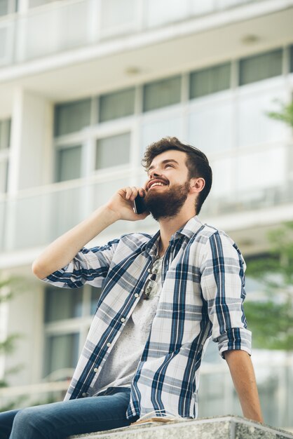 Vista de ángulo bajo de hipster barbudo hablando por teléfono fuera del edificio