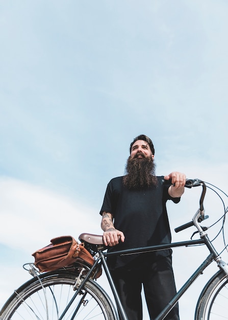 Vista de ángulo bajo de un hombre barbudo de pie con su bicicleta contra el cielo azul