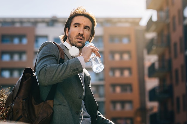 Vista de ángulo bajo del hombre de negocios bebiendo agua de una botella mientras está de pie en la ciudad y mirando hacia otro lado