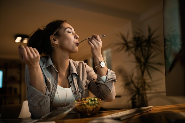 Vista de ángulo bajo de una mujer joven comiendo ensalada con los ojos cerrados mientras usa la computadora por la noche en casa