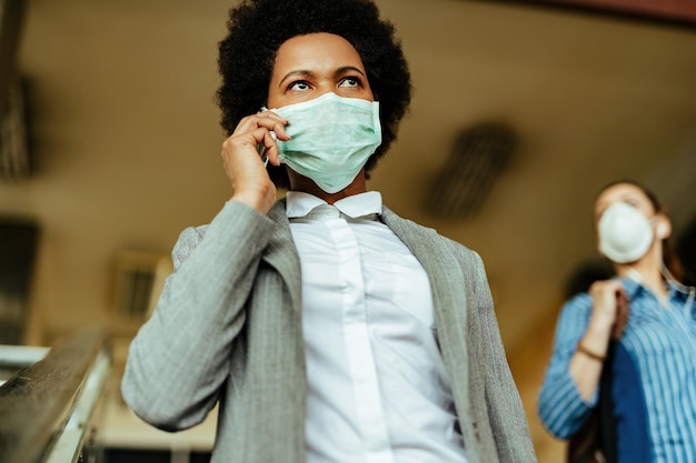 Vista de ángulo bajo de una mujer de negocios afroamericana caminando por un corredor público con máscara protectora y comunicándose por teléfono móvil