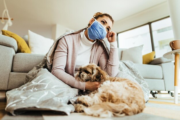 Vista de ángulo bajo de una mujer pensativa pensando mientras usa una máscara protectora y se sienta en el suelo con su perro en casa