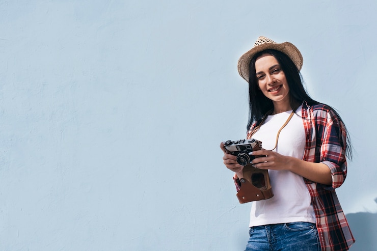 Foto gratuito vista de ángulo bajo de mujer sonriente con sombrero con cámara retro de pie contra el cielo