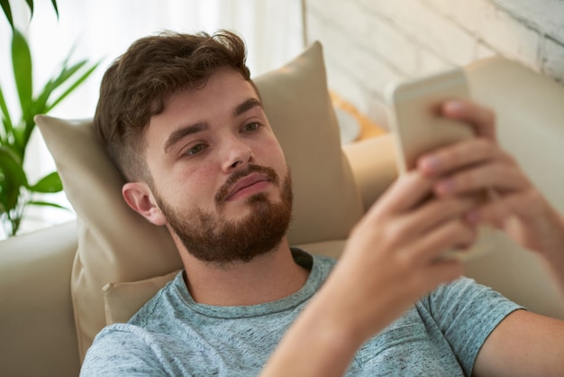 Vista de ángulo superior del chico joven que usa la aplicación móvil descansando en el sofá en casa