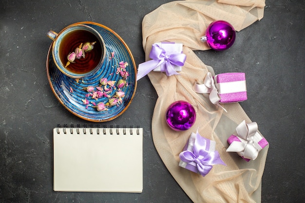 Vista anterior de accesorios de decoración de regalos coloridos para Navidad en una toalla de color nude y una taza de té junto al cuaderno sobre fondo negro