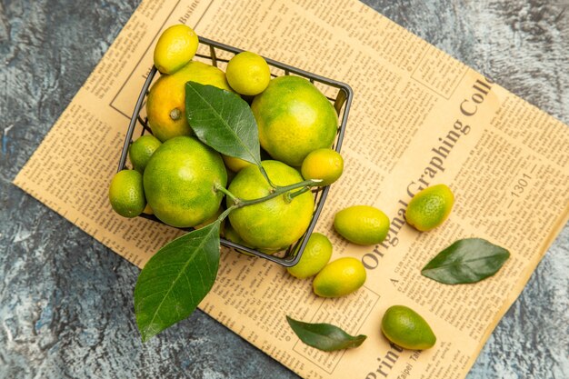 Vista anterior de la canasta negra con mandarinas verdes frescas y kumquats en periódicos sobre fondo gris