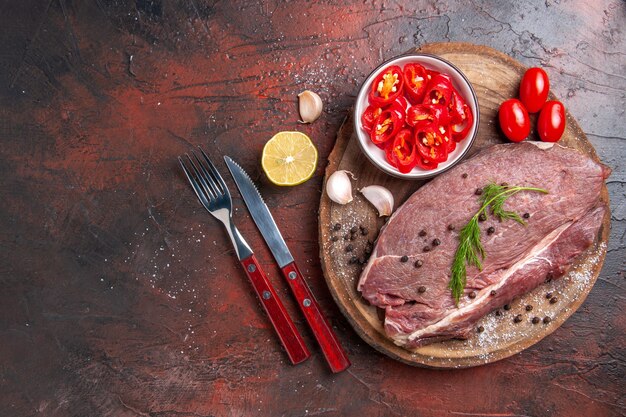 Vista anterior de la carne roja en la bandeja de madera y ajo verde pimiento picado botella de aceite caído tenedor y cuchillo en la mesa oscura