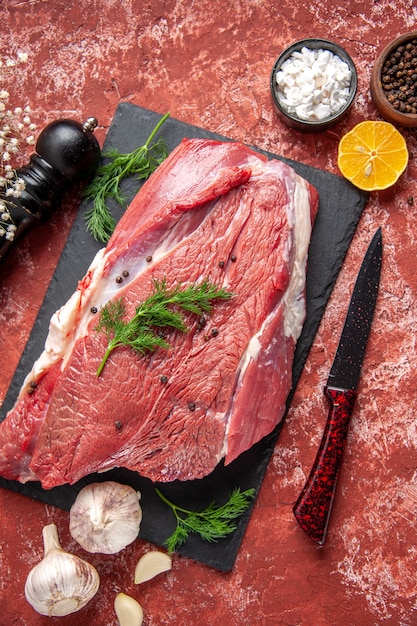 Vista anterior de la carne roja fresca cruda con verde y pimienta en el tablero negro cuchillo ajo especias limón martillo de madera limón sobre fondo rojo pastel de aceite