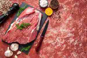 Foto gratuita vista anterior de carne roja fresca cruda con verde y pimienta en tablero negro cuchillo martillo de madera sal limón sobre fondo rojo pastel de aceite