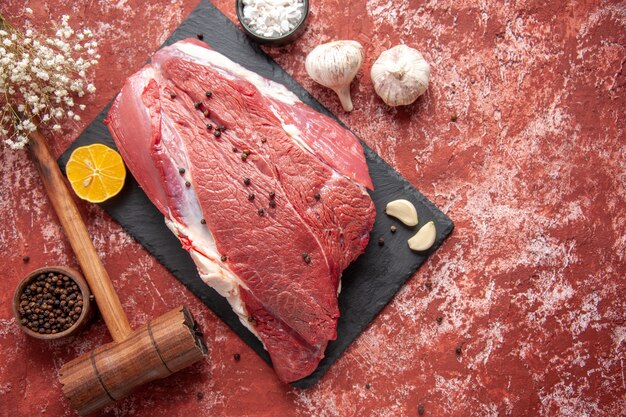 Vista anterior de carne roja fresca con pimienta en tablero negro cuchillo ajo especias limón martillo de madera marrón limón sobre fondo rojo pastel de aceite