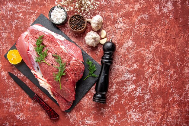 Vista anterior de carne roja fresca con verde y pimienta en tablero negro cuchillo ajo especias limón martillo de madera limón en el lado derecho sobre fondo rojo pastel al óleo