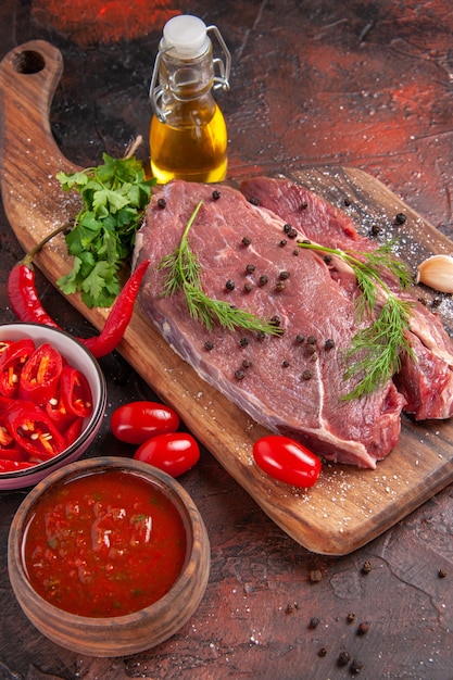 Vista anterior de la carne roja sobre la tabla de cortar de madera y ajo verde pimiento picado botella de aceite caído sobre fondo oscuro