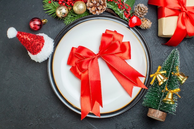 Vista anterior de la cinta roja en forma de arco en el plato de cena árbol de navidad ramas de abeto cono de coníferas caja de regalo sombrero de santa claus sobre fondo negro
