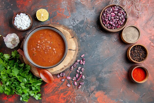 Vista anterior de la clásica sopa de tomate en un tazón azul cuchara en bandeja de madera, sal de ajo y limón, un manojo de verde en la tabla de colores mezclados