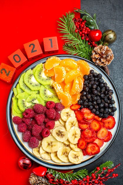 Vista anterior de la colección de frutas frescas en el plato de cena, accesorios de decoración, ramas de abeto y números, calcetín de navidad en una servilleta roja sobre fondo negro