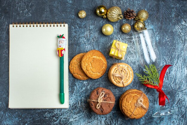 Vista anterior de la copa de vidrio caído con cinta roja y accesorios de decoración junto al cuaderno de galletas apiladas sobre fondo oscuro