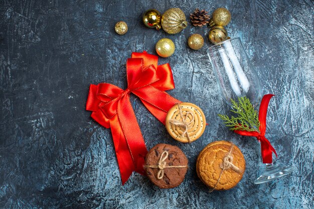 Vista anterior de la copa de vidrio caído con cinta roja y accesorios de decoración junto a galletas apiladas y cinta en forma de arco sobre fondo oscuro