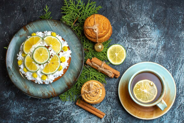 Vista anterior de cremoso pastel delicioso y ramas de abeto limón canela limas junto a una taza de té negro sobre fondo oscuro