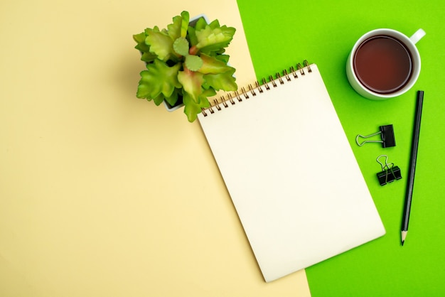 Vista anterior del cuaderno blanco con bolígrafo junto a una taza de té maceta sobre fondo blanco y amarillo