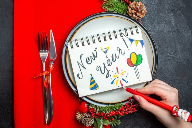 Vista anterior del cuaderno con escritura de año nuevo y dibujos en un plato con accesorios de decoración, ramas de abeto y cubiertos en una servilleta roja