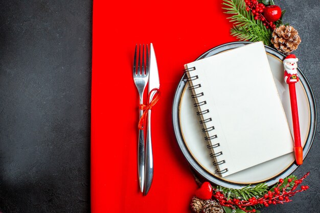 Vista anterior de un cuaderno de espiral y un bolígrafo en un plato con accesorios de decoración, ramas de abeto y cubiertos en una servilleta roja