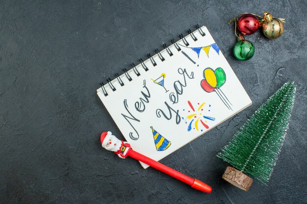 Vista anterior del cuaderno de espiral con escritura de año nuevo y bolígrafo junto a los accesorios de decoración del árbol de navidad sobre fondo oscuro