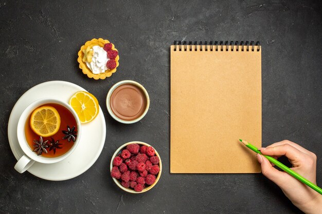 Vista anterior del cuaderno y una sabrosa cena con deliciosos panqueques frescos en un plato blanco y una taza de té negro, chocolate y miel de frambuesa sobre fondo oscuro