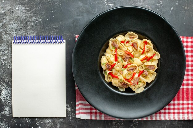 Vista anterior de la deliciosa conchiglie con verduras y verduras en un plato y un cuchillo sobre una toalla despojada de rojo y un cuaderno sobre fondo gris