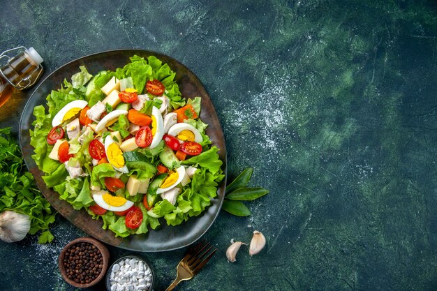 Vista anterior de deliciosa ensalada casera con muchos ingredientes en un plato y especias tenedor de botella de aceite caído en mesa de mezcla de colores verde negro con espacio libre