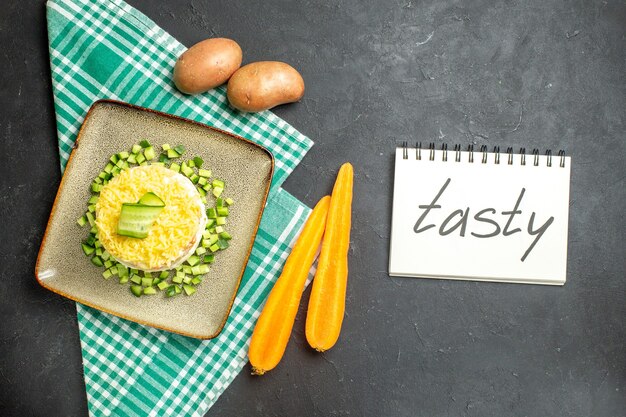 Vista anterior de una deliciosa ensalada servida con pepino picado en zanahorias y papas de toalla verde a la mitad dobladas junto al cuaderno con deliciosas incsripciones sobre fondo oscuro