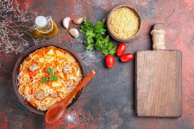 Vista anterior de una deliciosa sopa de fideos con pollo y pasta cruda en un pequeño tazón marrón y una cuchara de ajo, tomates y verduras, tabla de cortar sobre el fondo oscuro
