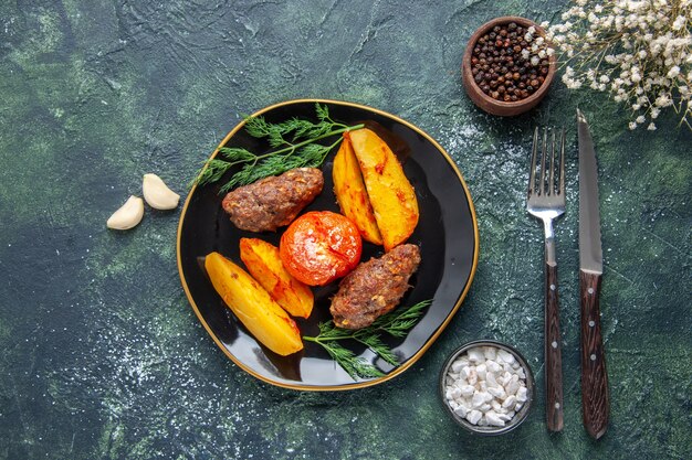 Vista anterior de deliciosas chuletas de carne al horno con patatas y tomates en una placa negra cubiertos de especias de ajo en verde negro mezcla de colores de fondo