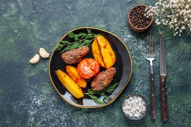 Vista anterior de deliciosas chuletas de carne al horno con patatas y tomates en una placa negra cubiertos de especias de ajo en verde negro mezcla de colores de fondo