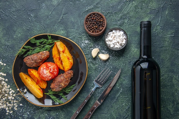 Vista anterior de deliciosas chuletas de carne al horno con patatas y tomates en una placa negra, especias, ajo, botella de vino sobre fondo verde negro mezcla de colores