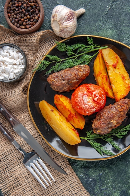 Vista anterior de deliciosas chuletas de carne al horno con patatas y tomates en una placa negra, especias, ajos, cubiertos en verde negro mezcla de colores de fondo