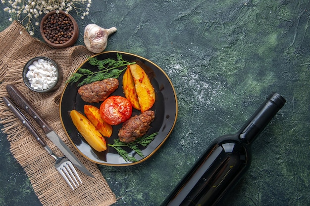 Vista anterior de deliciosas chuletas de carne al horno con patatas y tomates en una placa negra, especias, ajos, cubiertos, vino, sobre fondo verde negro mezcla de colores