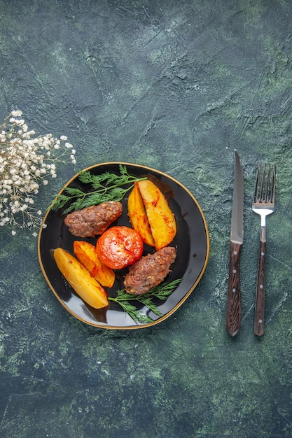 Vista anterior de deliciosas chuletas de carne al horno con patatas y tomates en un plato negro cubiertos con flores blancas sobre fondo verde negro de colores mezclados