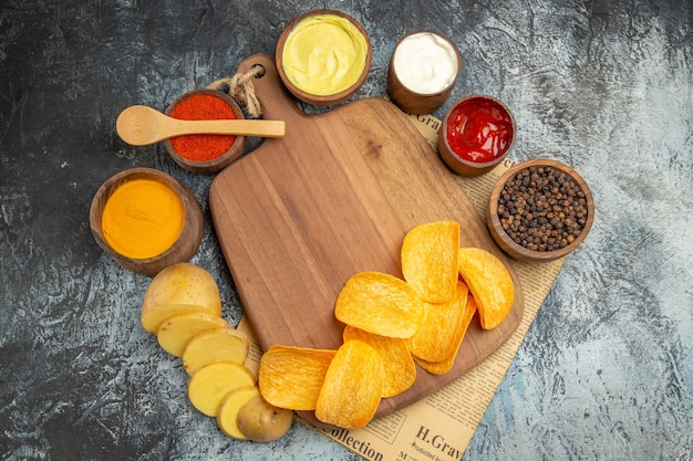 Vista anterior de deliciosas papas fritas caseras en la tabla de cortar de madera diferentes especias y sabores en el periódico sobre la mesa gris