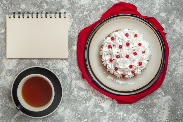 Vista anterior del delicioso pastel cremoso decorado con frutas sobre una toalla roja y una taza de té negro junto al portátil sobre fondo de hielo