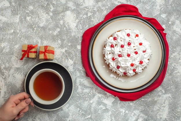 Vista anterior de un delicioso pastel cremoso decorado con frutas en una toalla roja mano sosteniendo una taza de té negro pequeñas cajas de regalo sobre fondo de hielo