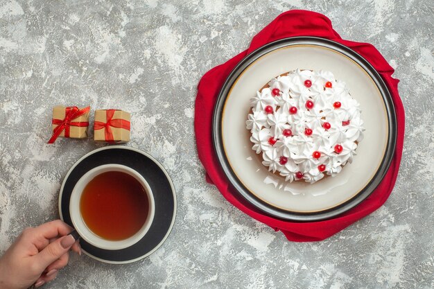 Vista anterior de un delicioso pastel cremoso decorado con frutas en una toalla roja mano sosteniendo una taza de té negro pequeñas cajas de regalo sobre fondo de hielo