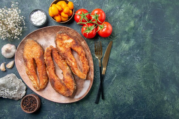 Vista anterior de un delicioso pescado frito en una placa marrón y cubiertos, especias, alimentos en una mesa de colores mezclados con espacio libre