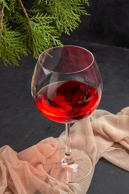 Vista anterior de delicioso vino tinto en una copa de cristal sobre una toalla y ramas de abeto sobre un fondo oscuro