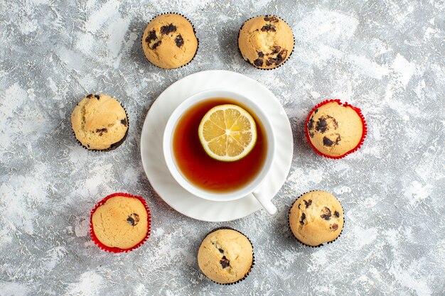 Vista anterior de deliciosos cupcakes pequeños con chocolate alrededor de una taza de té negro sobre la superficie del hielo