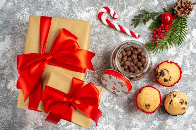 Vista anterior de deliciosos cupcakes pequeños y chocolate en una olla de vidrio y ramas de abeto junto al regalo con cinta roja sobre la superficie del hielo