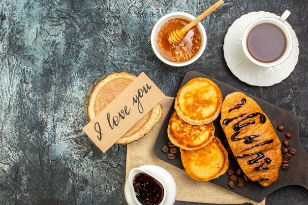 Vista anterior de deliciosos panqueques croissant sobre tabla de cortar de madera una taza de miel de té negro sobre superficie oscura
