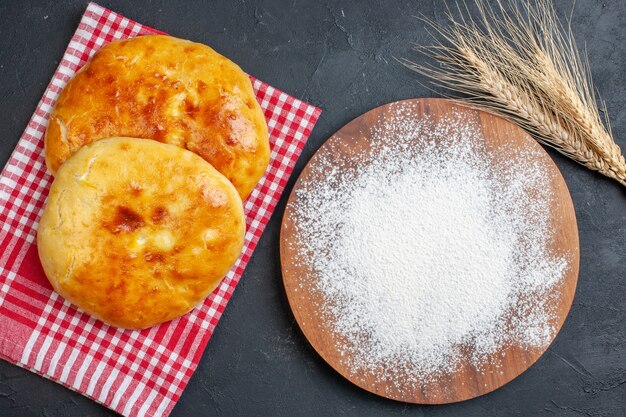 Vista anterior de deliciosos pasteles recién horneados en una toalla despojada de rojo y harina en la tabla de cortar de madera en blackground oscuro