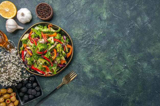 Vista anterior de ensalada vegana en un plato y tenedor de ajo flor blanca botella de aceite caído de oliva sobre fondo oscuro