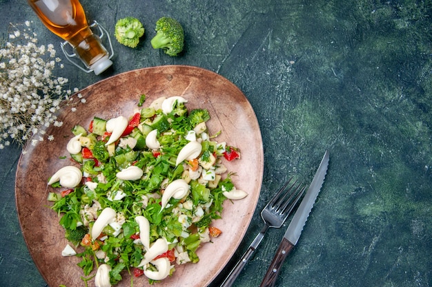 Vista anterior ensalada de verduras frescas dentro de un elegante plato con cubiertos sobre fondo azul oscuro